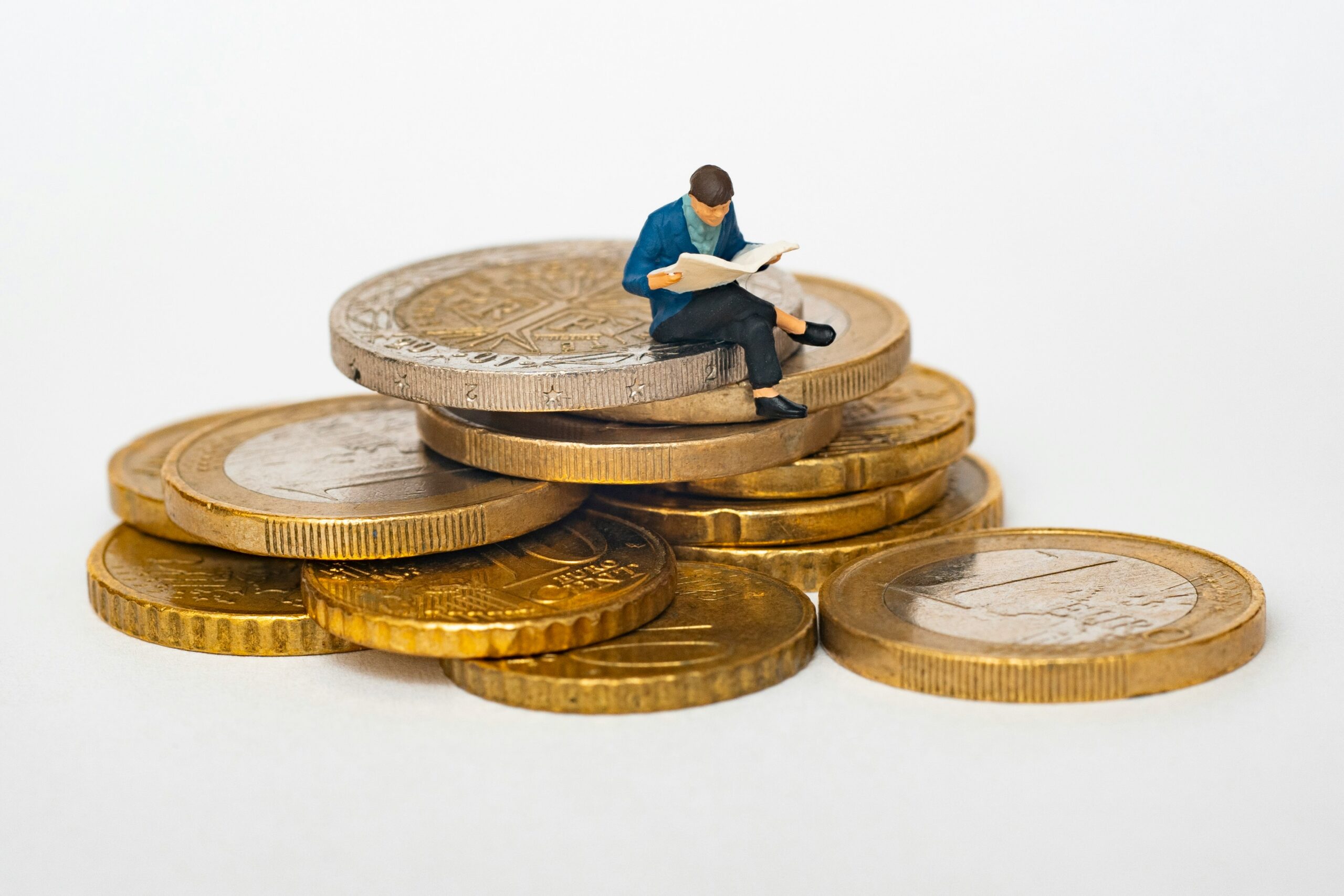 Miniatuurfiguurtje in pak leest een krant, zittend op een stapel euromunten